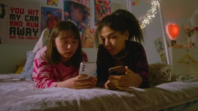 Two multicultural best friends in the cozy bedroom talk and take funny selfie pictures using mobile phone. Happy teenage girls spend leisure time at home lying on the bed. Besties having fun together.