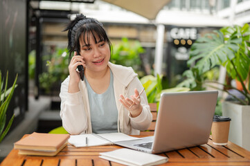 A young Asian woman is working remotely at an outdoor space, talking on the phone with her client.