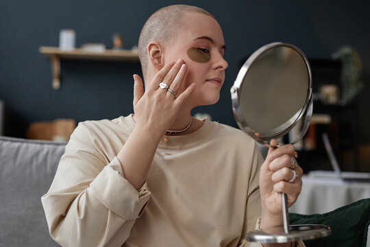 Portrait of young Caucasian woman with undereye patches on face holding mirror applying nourishing cream on skin