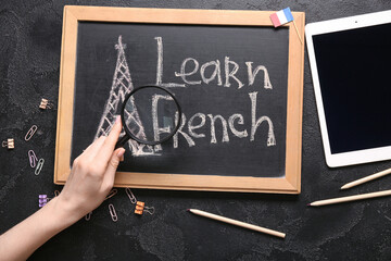 Female hand, chalkboard with text LEARN FRENCH, stationery and tablet computer on dark background,...