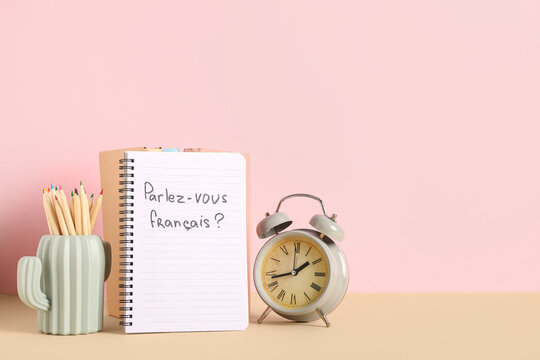 Notebook with French phrase, pencils and alarm clock on table against color background