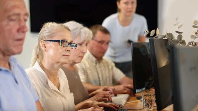 Motivated elderly woman in glasses mastering computer skills during group course with mentor standing on blurred background. Concept of curiosity of seniors towards technology