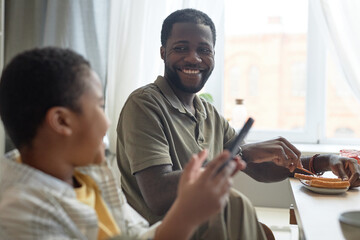 Portrait of smiling African American father looking at son using smartphone at kitchen table during breakfast