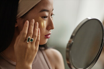 Closeup of young Asian woman holding mirror applying collagen patches under eyes