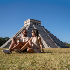 Lovely couple in love visiting Chichen Itza, sitting on the grass with the pyramid in the...