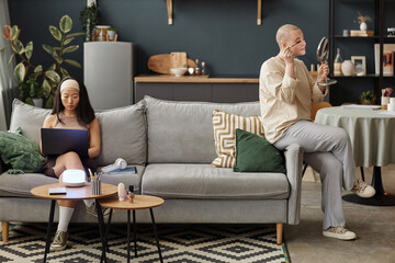 Wide shot of two diverse women spending time at home sitting on sofa in living room surfing...