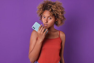 Young scared curly African American woman student feels fear after reading SMS message on phone and looks at camera with serious emotions in need of protection stands on purple background.