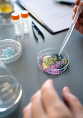 Close up of female chemist hands adding drop of liquid from from pipette over petri dish with mix of colorful glitters in environment research laboratory. Dangers in microplastics composition concept.
