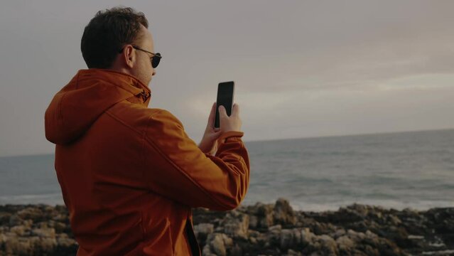 Back view on man hold smartphone and take picture of beautiful seascape. Young male walking on ocean coast and shooting video of nature using cellphone camera