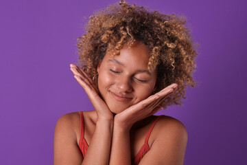 Young positive ethnic African American woman teen puts palm under chin and smiles closing eyes with pleasure or good memories of vacations spent with friends stands posing on purple background.