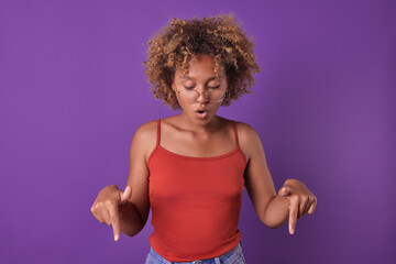 Young surprised casual African American woman teenager points finger down paying attention to shocking offer to connect services with big discount stands on isolated purple background. Copy space