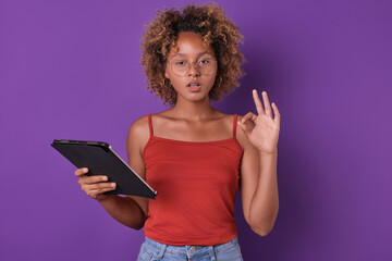 Young beautiful African American woman freelancer with electronic tablet shows OK gesture and looks at camera reporting completion of order or successful completion of project stands in purple studio.