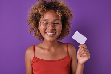 Young happy beauty African American woman teenager with smile demonstrates plastic bank card with possibility of online payment and large credit limit of stands on isolated purple background.