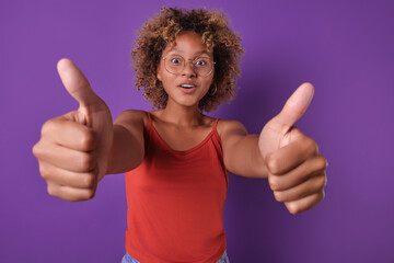 Young inspired casual African American woman teen shows two hands thumbs up expressing approval and agreement or confirmation of excellent quality of purchased product stands posing in purple studio.