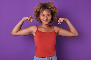 Young happy casual African American woman zoomer showing biceps taking strongman pose and communicating desire to be strong and independent from men stands on purple studio background.