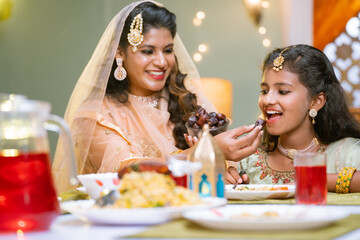 Happy indian mother feeding dates to daughter during ramadan iftar dinner at home - concept of...