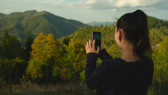 Young woman making photos of mountains on mobile phone device. Girl standing on top of mountain peak and photographing scenic landscape on cellphone camera