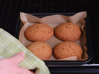 A woman taking a baking tray with four oatmeal cookies on it out of an oven.