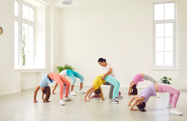 Fototapeta premium Kids doing gymnastics. Group of children doing gymnastic exercises at a sports school. Female trainer helps little flexible sporty gymnast girls who are doing a bridge position all together