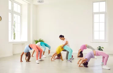 Fototapete Kids doing gymnastics. Group of children doing gymnastic exercises at a sports school. Female trainer helps little flexible sporty gymnast girls who are doing a bridge position all together © Studio Romantic