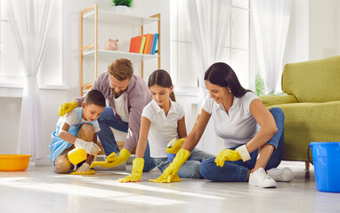 Happy smiling young parents with two children boy and girl cleaning floor together with rags and detergent in the living room at home. Family housework, housekeeping and household concept.