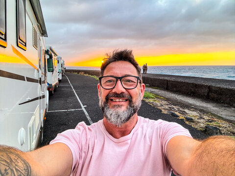 One happy man taking selfie picture on the side of a modern camper van motorhome. Traveler vanlife lifestyle people sharing on social media adventure and journey. Amazing sunset in background. Parking