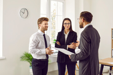 Corporate business team discussing something at a work meeting. Group of three people talking about work. Man and woman standing in the office and listening to their manager - 772887302