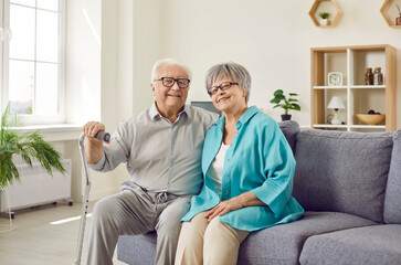Portrait of a happy senior family couple wife and husband with crutch sitting on sofa in the living room at home. Two smiling gray-haired retired man and woman enjoying time together on retirement. - 772889935