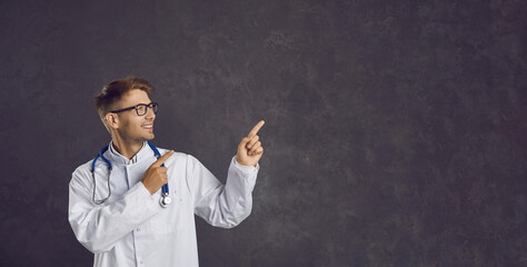 Side view of happy young male nurse or doctor in white lab coat smiling and pointing his index fingers at blank grey advertising copy space banner background, presenting new drug or medical product