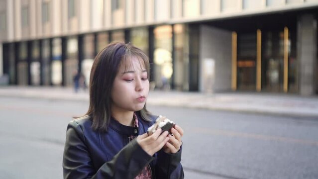 Slow-motion video of an Okinawan woman in her 20s in winter clothes eating street food pork egg rice balls while walking on Kokusai Street in Naha City, Okinawa Prefecture 沖縄県那覇市の国際通りを歩きながらストリートフードのポー