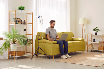 Young man sitting on sofa and working with laptop in the living room at home while receiving IV...