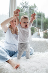 A 5-month-old baby and his  beautiful blonde mom relax, play and laugh in bed in the bedroom. People a dressed in light home clothes, the family chooses natural textiles