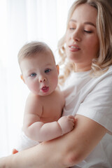 A 5-month-old baby and his  beautiful blonde mom relax, play and laugh in bed in the bedroom. People a dressed in light home clothes, the family chooses natural textiles
