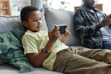 Portrait of teenage African American boy using smartphone and watching videos online while relaxing on couch at home