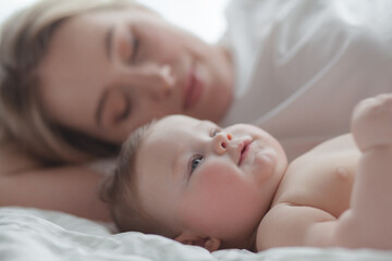 A 5-month-old baby and his  beautiful blonde mom relax, play and laugh in bed in the bedroom. People a dressed in light home clothes, the family chooses natural textiles