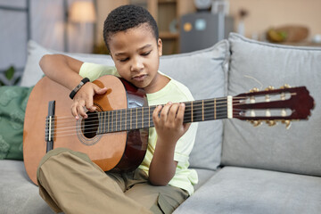 Front view portrait of young Black boy learning to play acoustic guitar sitting on couch at home...