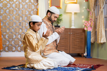 Indian Muslim father with kid doing namaz or praying during ramadan festival celebration at home -...