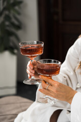 Two young women in bathrobes drink cocktails and pose in the bedroom. Women's party concept