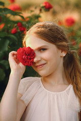 Young girl with blonde hair holding a red rose flower near her face while posing in the rose garden in summer. The charm of youth