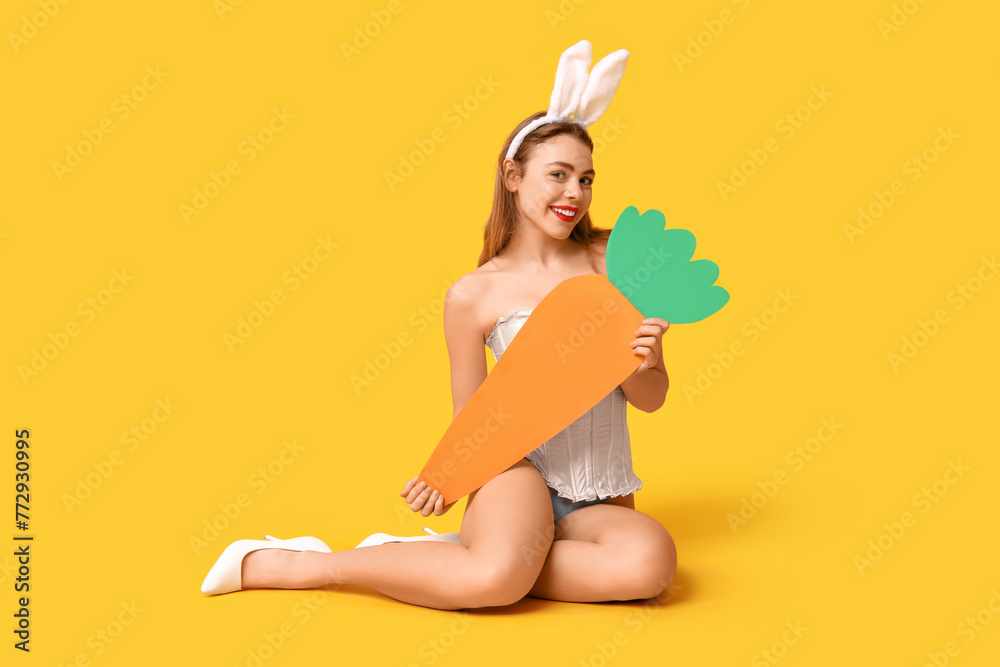 Wall mural sexy smiling young woman in bunny ears with paper carrot sitting on yellow background. easter celebr