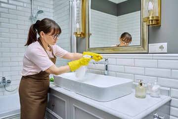 Woman in apron with detergent spray washcloth cleaning bathroom