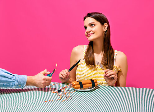 Smiling young woman sitting at table with player and sausages imitating explosive item against pink background. Surrealism. Bang. Concept of food pop art photography, creativity, quirky style