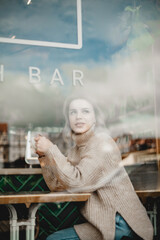 Woman in Cafe Gazing Out Window. A woman sits at a cafe table, dressed in a cozy sweater and jeans, lost in thought as she gazes out the window, capturing a moment of contemplation and relaxation