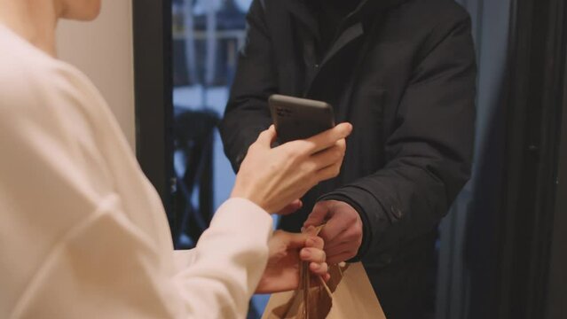 Slow motion of mature Caucasian woman standing in front of young man delivering food products to her, accepting delivery status on smartphone