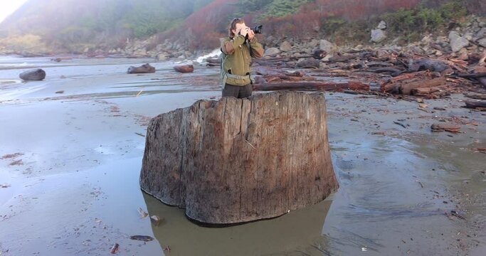A man is standing on a log in the ocean. He is taking a picture of the beach.