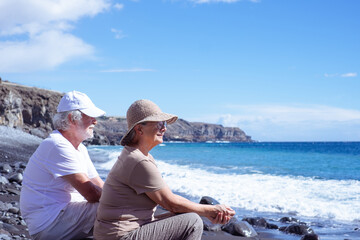 Relaxed senior couple in vacation sitting on the seashore looking waves splashing on the beach enjoying retirement lifestyle and freedom, horizon over sea