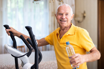Senior man with bottle of water using elliptical trainer at home