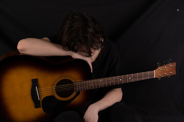 Male teenager with acoustic guitar on black background. With long hair in black clothes