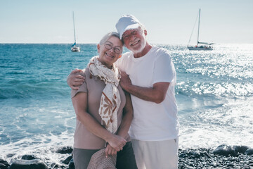 Portrait of smiling senior couple in vacation standing on the seashore looking at camera enjoying...