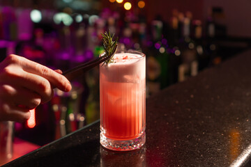 Close-up of the bartender adorns a glass with a sprig of rosemary with a strawberry cocktail, ice and foam while standing at the bar. Bartender prepares a cocktail